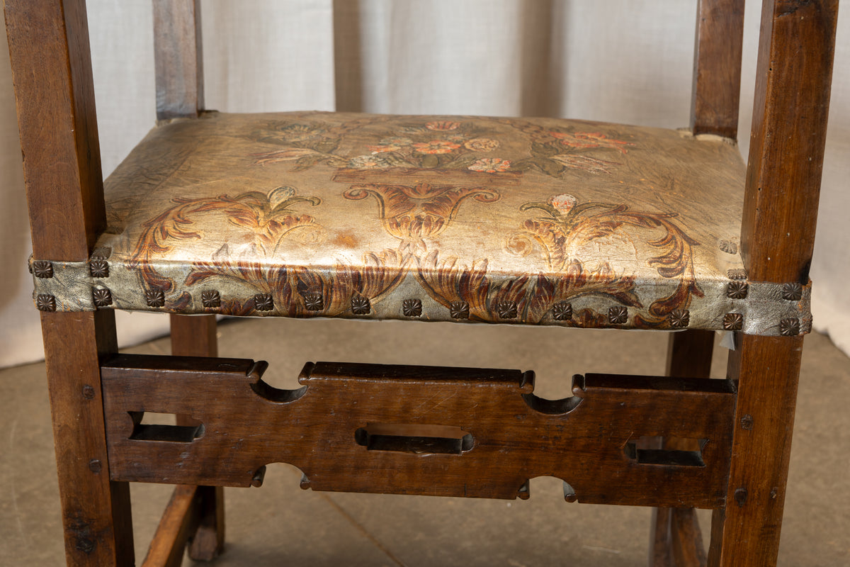 A close-up of an antique wooden chair featuring a distressed, patterned fabric seat cushion with floral motifs in brown, gold, and muted red, secured by metal brackets, set against a neutral draped backdrop.