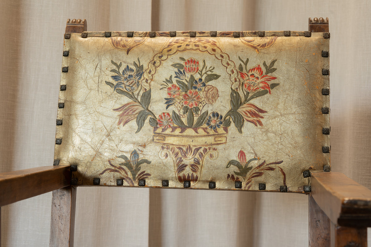 A close-up of an antique upholstered chair backrest featuring a floral tapestry design on aged beige fabric, bordered by dark metal studs, set against a neutral curtain background.