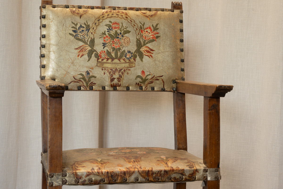 An antique wooden armchair with a floral tapestry backrest and seat cushion, featuring brass nailhead trim, set against a neutral draped fabric backdrop.