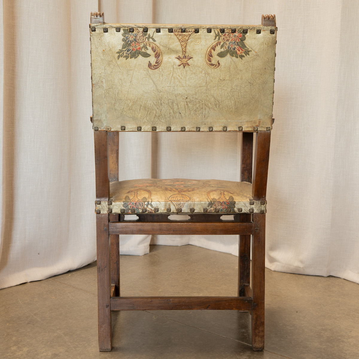 An antique wooden dining chair with a distressed beige upholstered backrest and seat featuring floral embroidery and brass nailhead trim, set against a draped cream fabric backdrop.