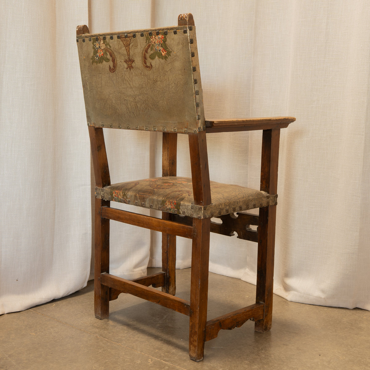 An antique wooden chair with a distressed beige upholstered seat and backrest, featuring floral embroidery on the back panel and brass nailhead trim, set against a draped white fabric backdrop.
