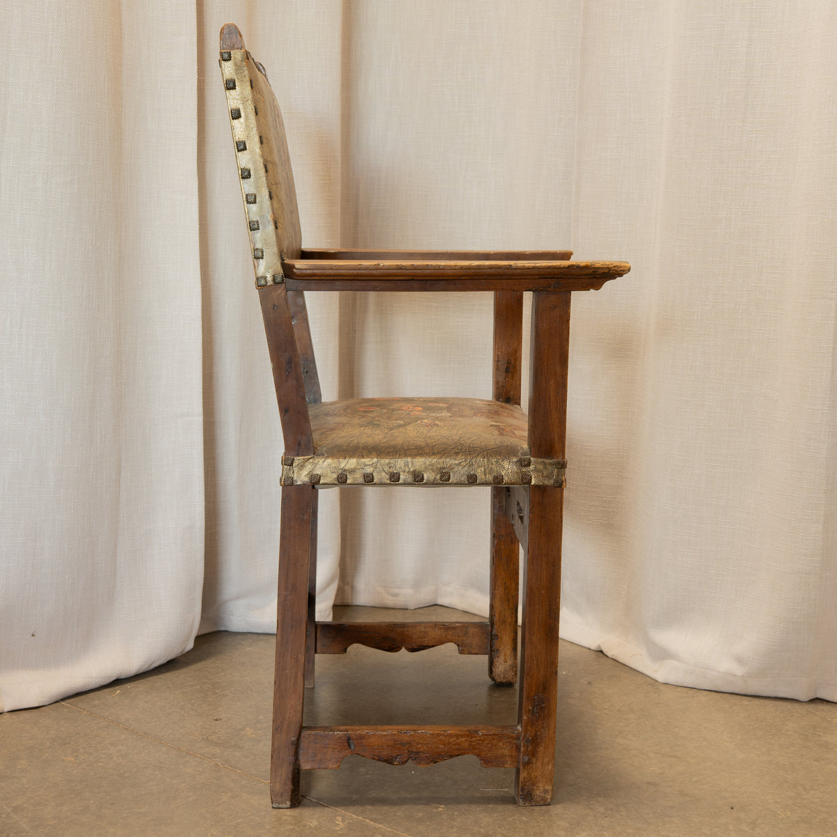 An antique wooden armchair with a distressed brown finish, a worn beige upholstered seat and backrest, and brass nailhead trim, set against a draped cream fabric backdrop.