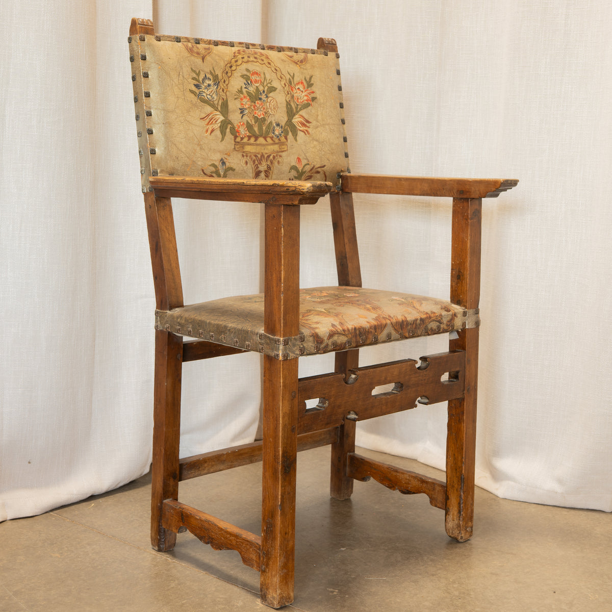 A vintage wooden armchair with a floral tapestry upholstered backrest and seat cushion, set against a draped white fabric backdrop.