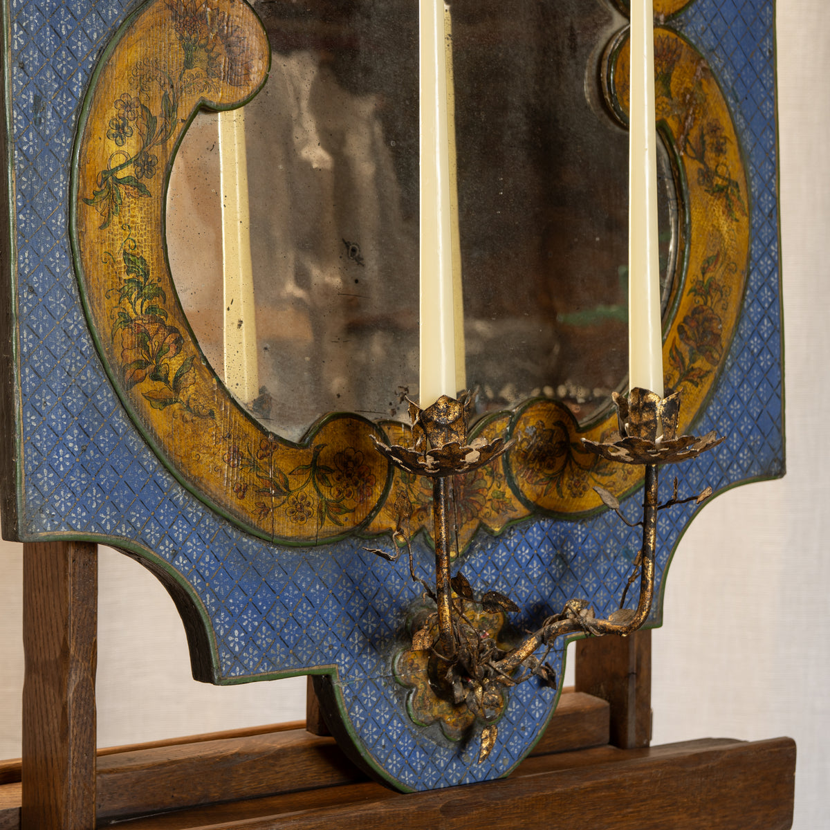 An antique decorative wall mirror with a blue patterned frame, gold floral border, and a metal candlestick-style support holding two cream candles.
