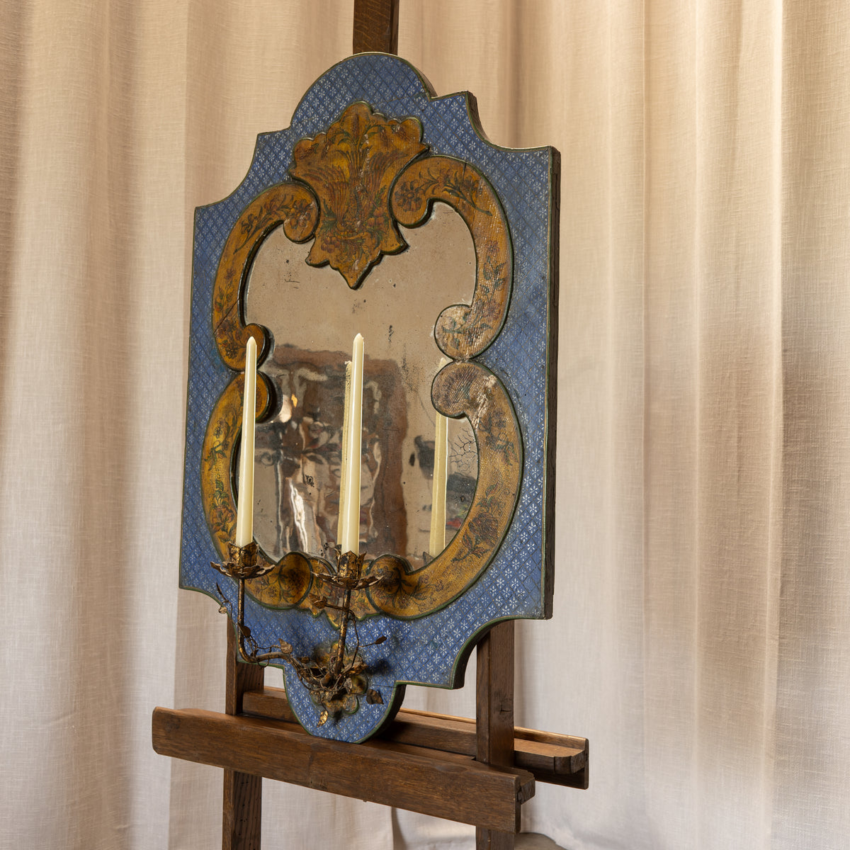 An ornate antique-style wall mirror with a blue patterned border and gold-toned scrollwork, mounted on a wooden easel against a beige curtain backdrop.