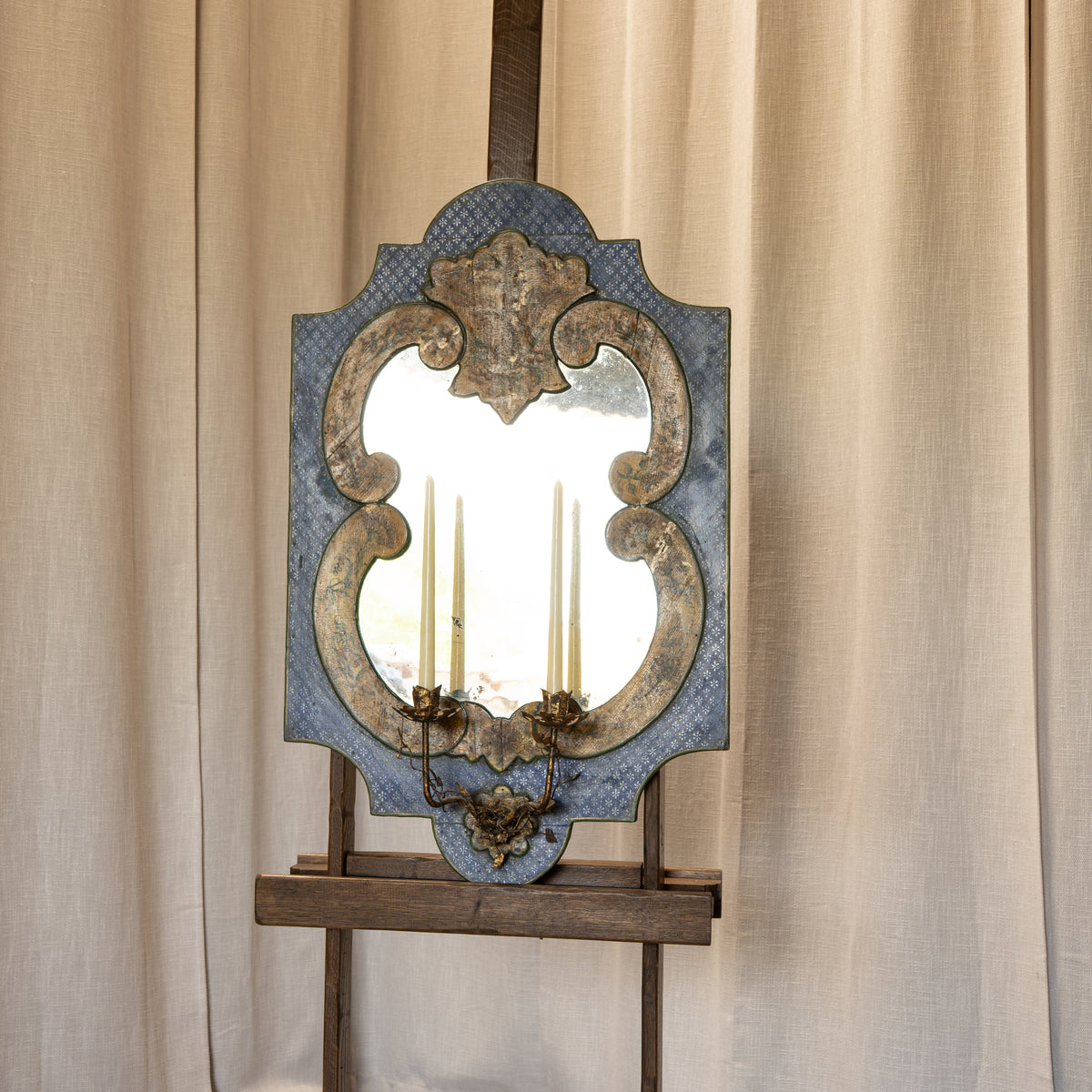 An ornate antique-style wall mirror with a blue patterned frame, gold-toned scrollwork, and built-in candle-style sconces, displayed on a wooden easel against beige curtains.