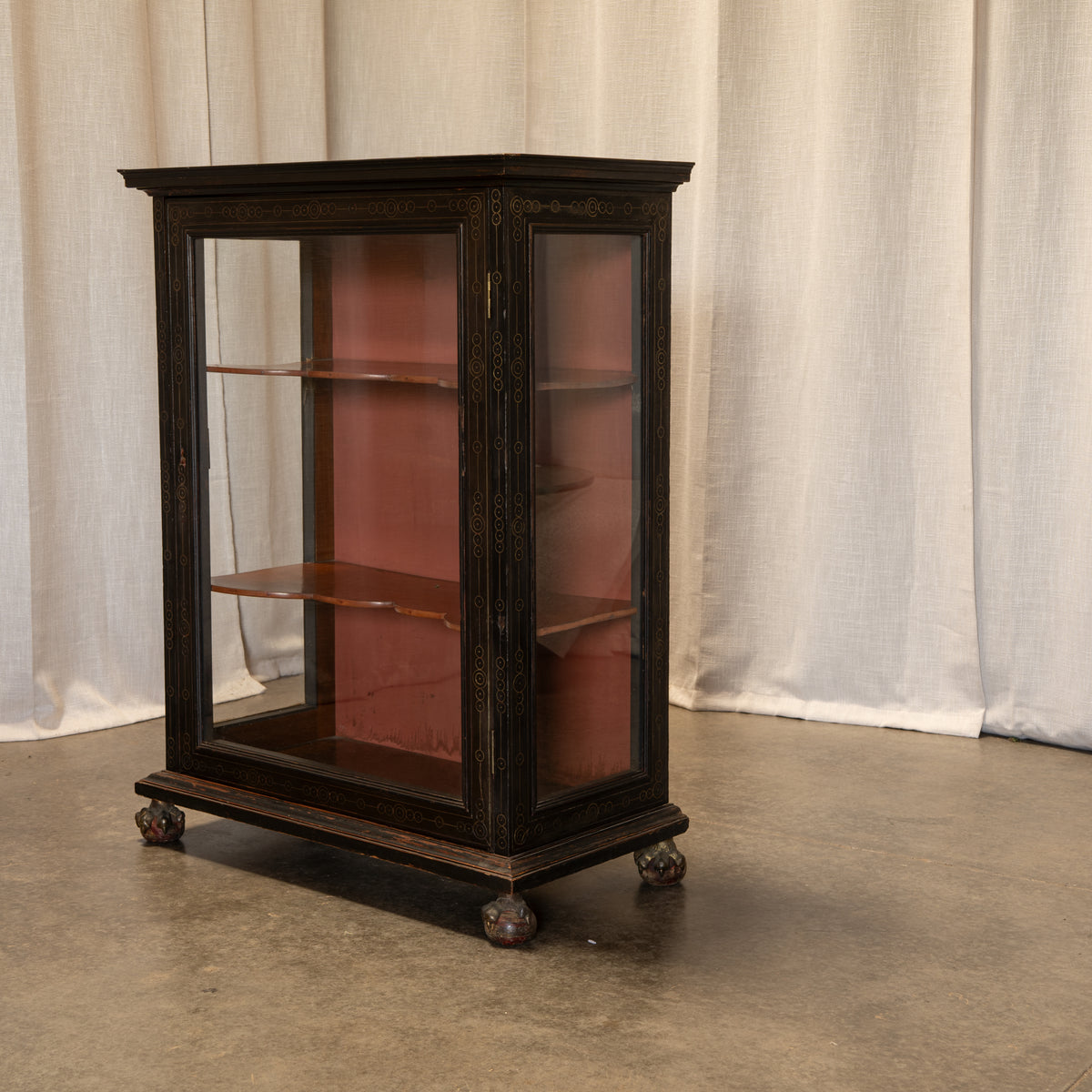 An antique dark wood display cabinet with glass doors and three interior shelves, set against a backdrop of draped white fabric and a concrete floor.