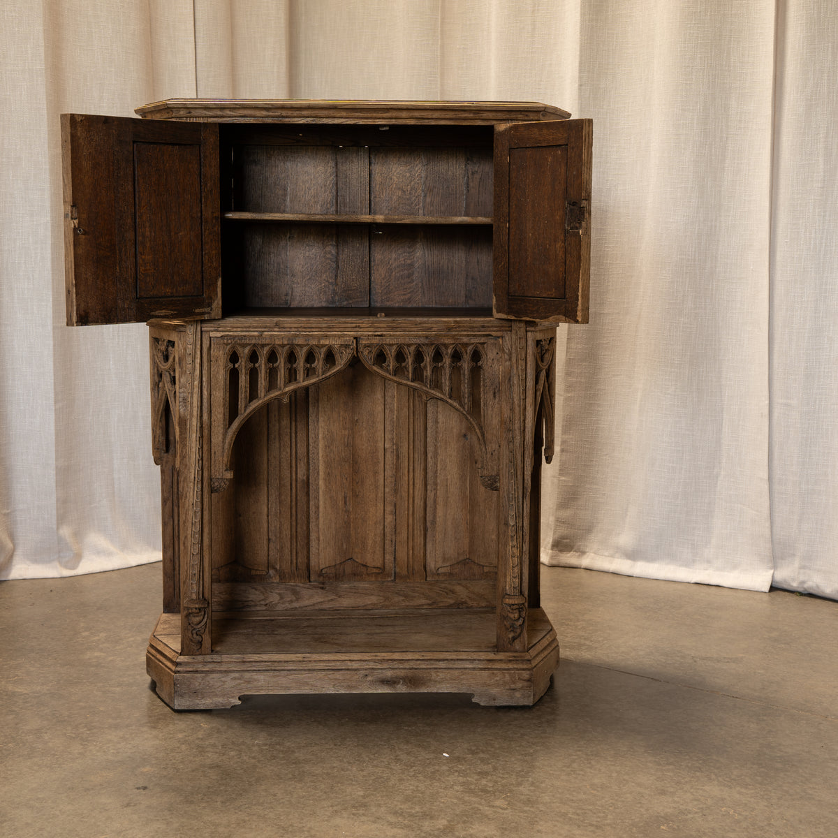 An antique carved wooden cabinet with open doors, displayed against a draped beige fabric backdrop on a concrete floor.