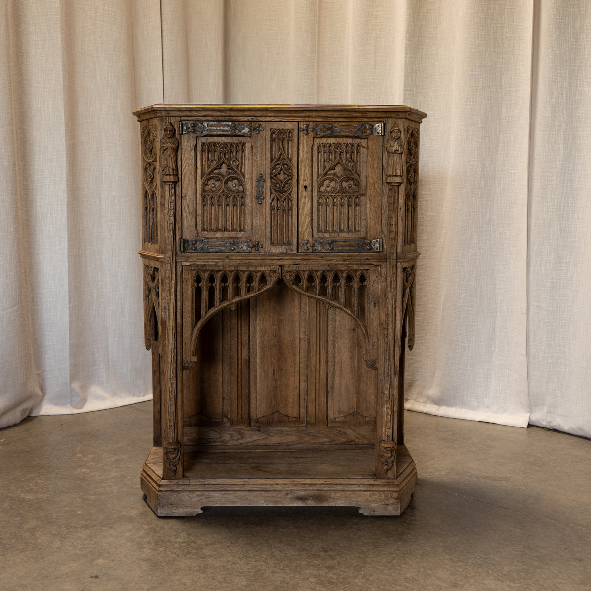 An ornate, vintage-style carved wooden cabinet or side table with Gothic arch details, photographed against a neutral beige fabric curtain backdrop.