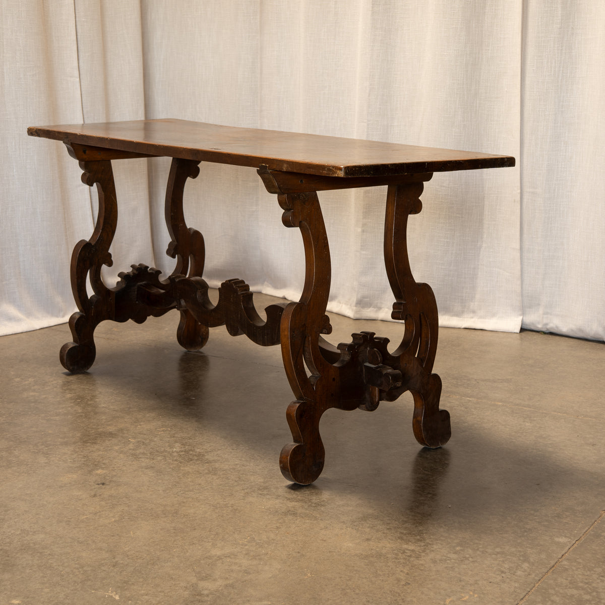An ornate dark brown wooden console table with carved legs and a rectangular top, set against a draped white fabric backdrop on a concrete floor.