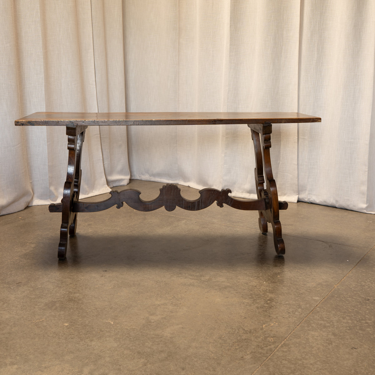 A dark wood console table with a rectangular top and ornate carved legs, set against a neutral beige curtain backdrop.