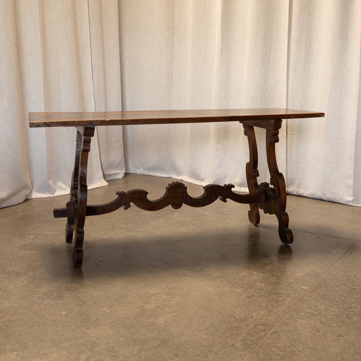 An antique dark wood console table with turned legs and a decorative center stretcher, set against a draped white fabric backdrop on a concrete floor.