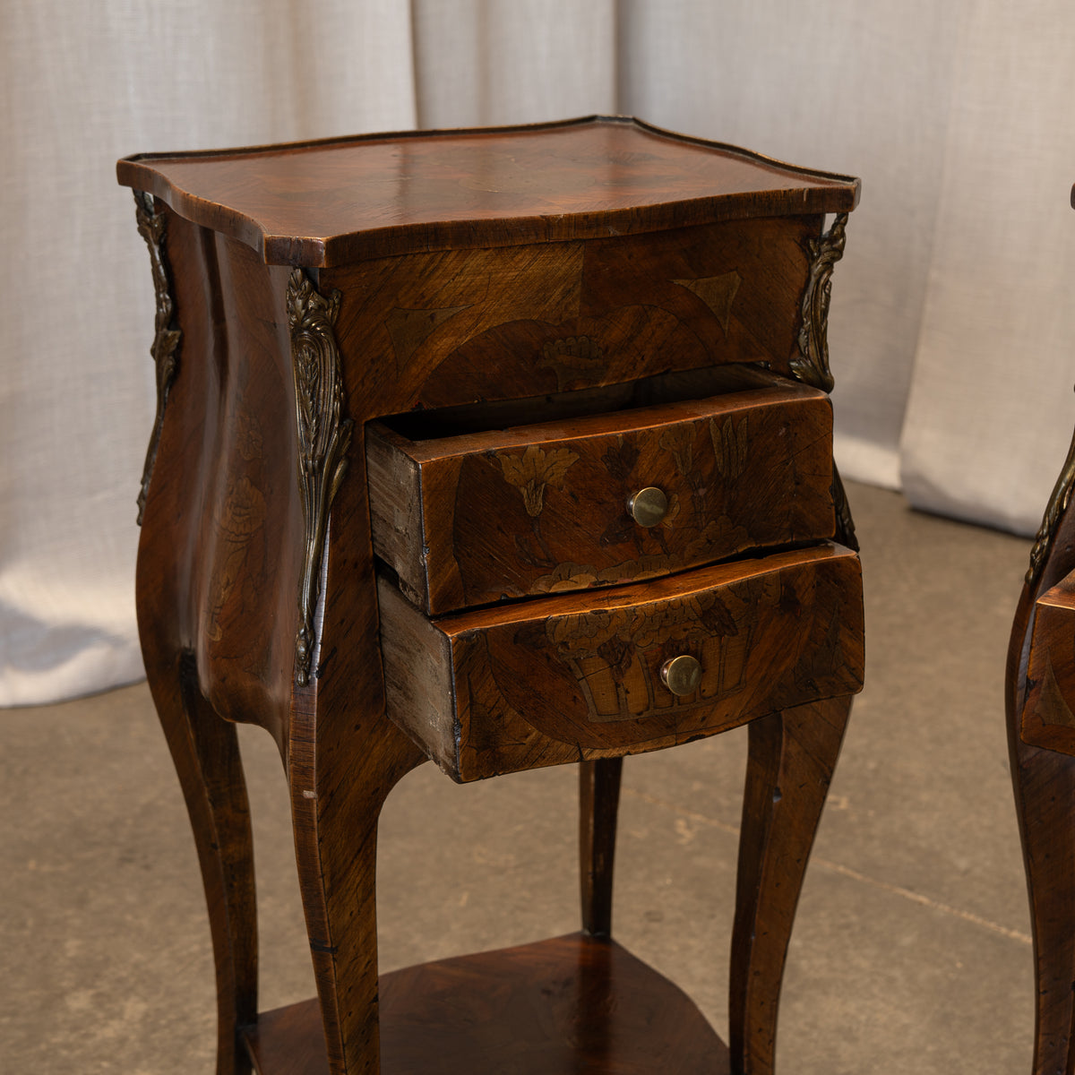 A vintage wooden nightstand with two drawers, featuring a rich dark brown wood grain and ornate metal drawer pulls, set against a neutral draped fabric backdrop.