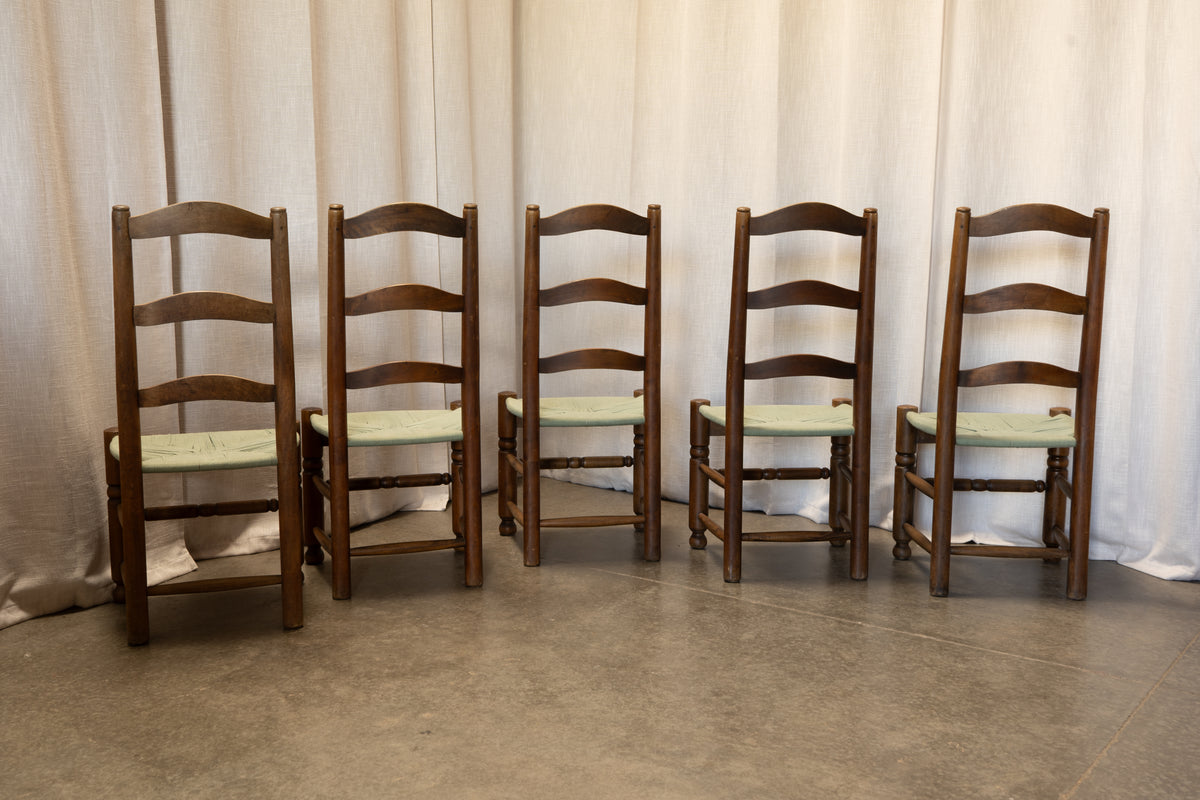 Five wooden ladder-back dining chairs with light green upholstered seats, lined up against a draped beige fabric backdrop on a concrete floor.
