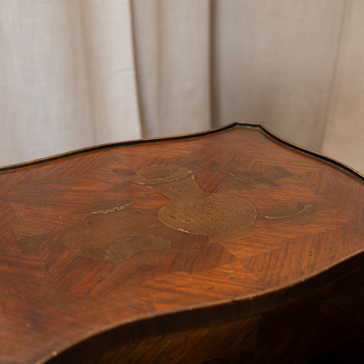 Close-up of a vintage wooden table top featuring a detailed geometric inlay pattern in warm brown tones, set against a neutral beige curtain background.