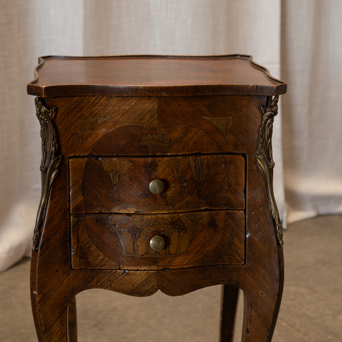 An antique wooden nightstand with two drawers, featuring intricate marquetry inlays, carved side supports, and round metal knobs, set against a neutral draped fabric background.