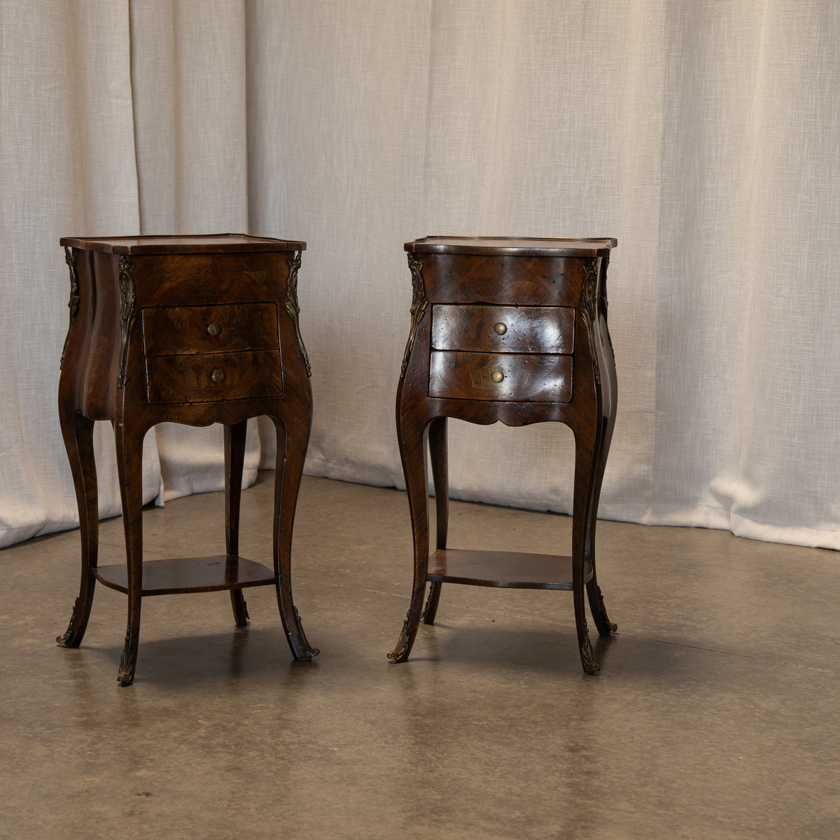 A pair of antique dark brown wooden nightstands with curved legs, single drawers, and lower shelves, set against a draped beige fabric backdrop.