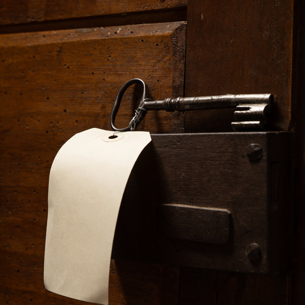 A vintage metal key hanging from a dark wooden door lock, with a blank white paper tag attached.