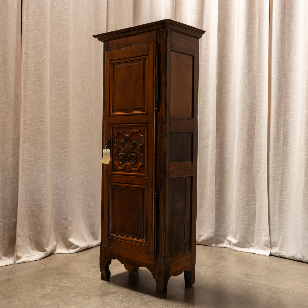 A tall, narrow dark brown wooden cabinet with carved panel detailing on the door, photographed indoors against draped beige curtains and a concrete floor.