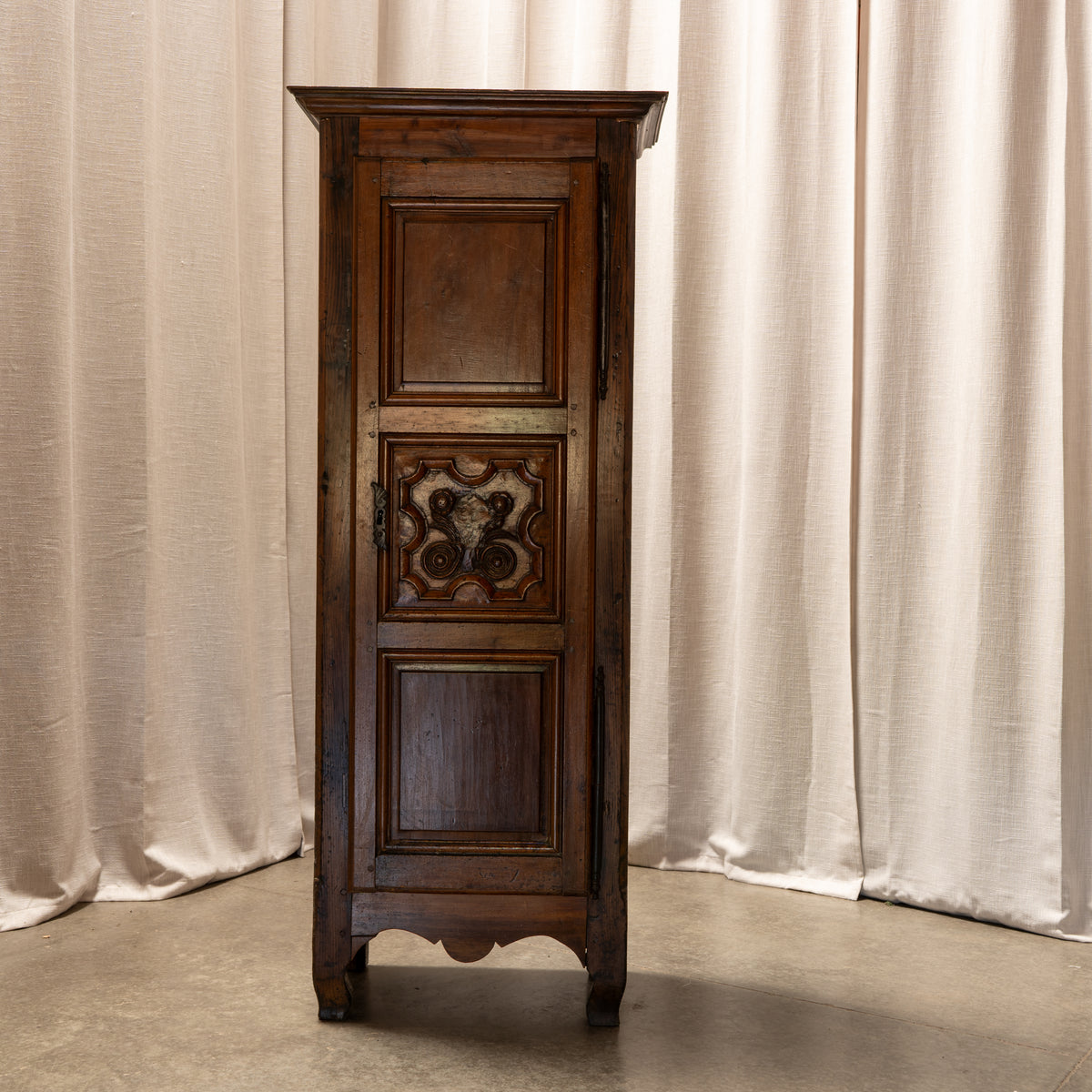A tall, narrow rustic dark brown wooden cabinet with carved details on the center door, set against a backdrop of beige fabric curtains.