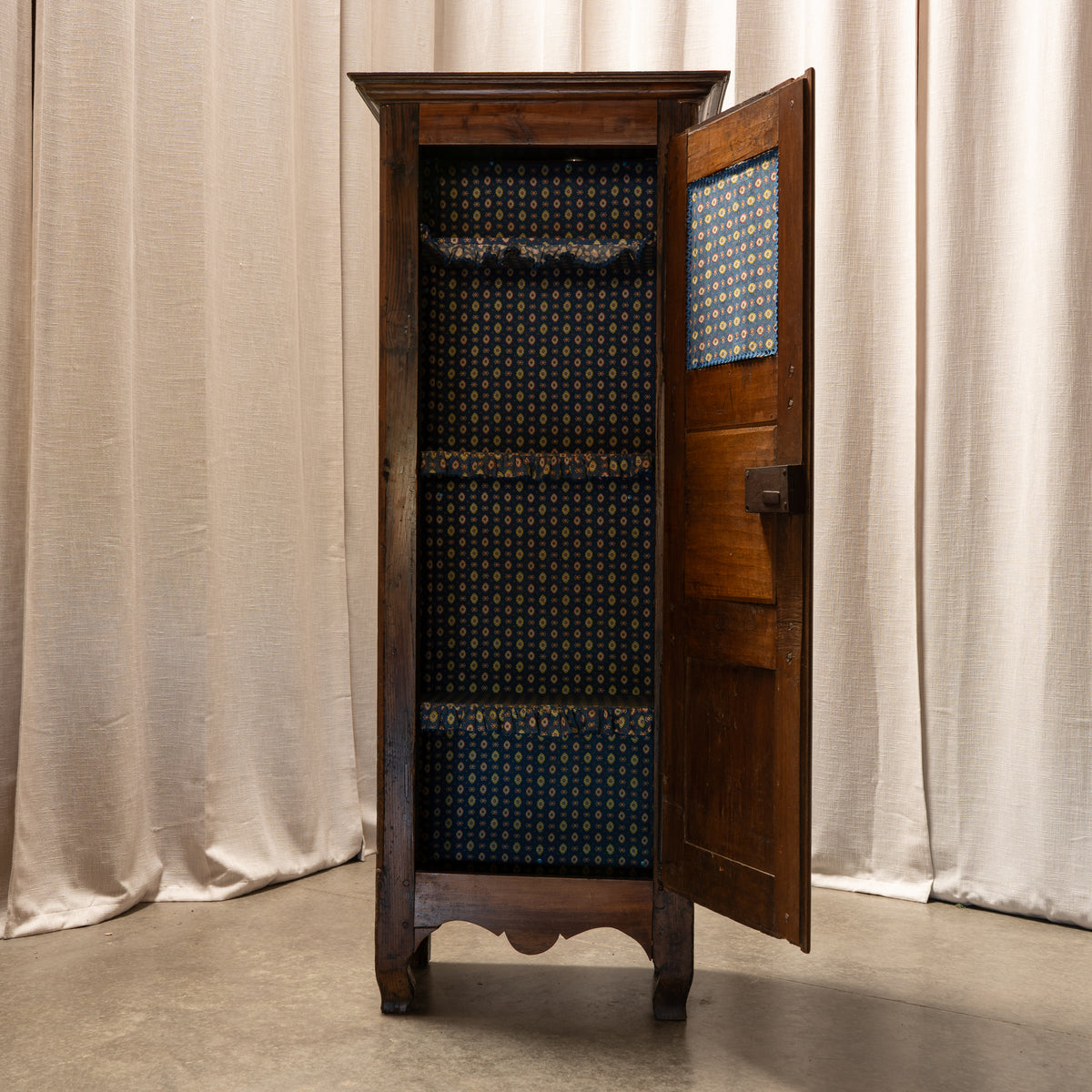 A tall vintage wooden display cabinet with one door open, showcasing dark blue patterned fabric lining and lace trim, set against draped beige curtains.