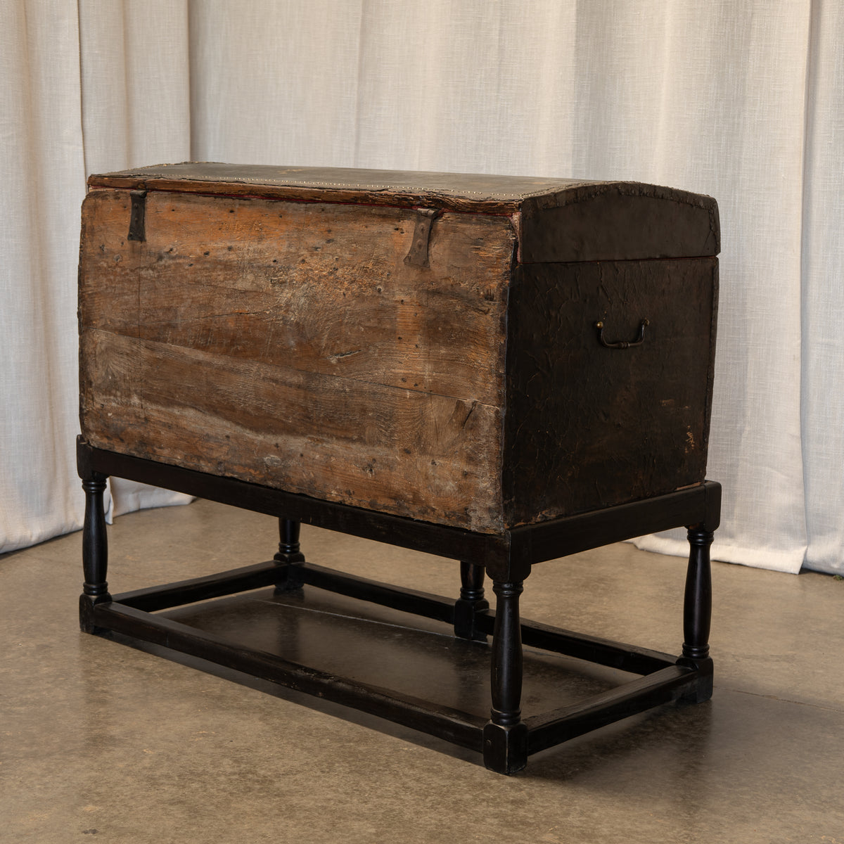 A rustic antique wooden chest with a dark brown painted exterior and a weathered natural wood front panel, resting on a black metal stand with turned legs, set against a draped white fabric backdrop.