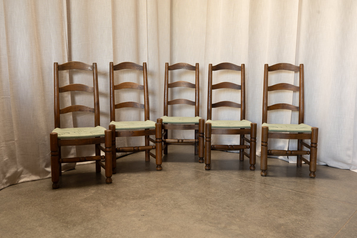 Six wooden ladder-back dining chairs with light green upholstered seats, arranged in a row against a beige fabric backdrop.