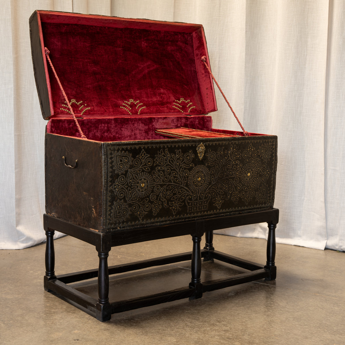 An antique dark brown wooden chest with intricate gold floral patterns, elevated on a black wooden stand. The chest is open, revealing a deep red velvet-lined interior with gold embroidery on the lid, set against a white curtain backdrop.