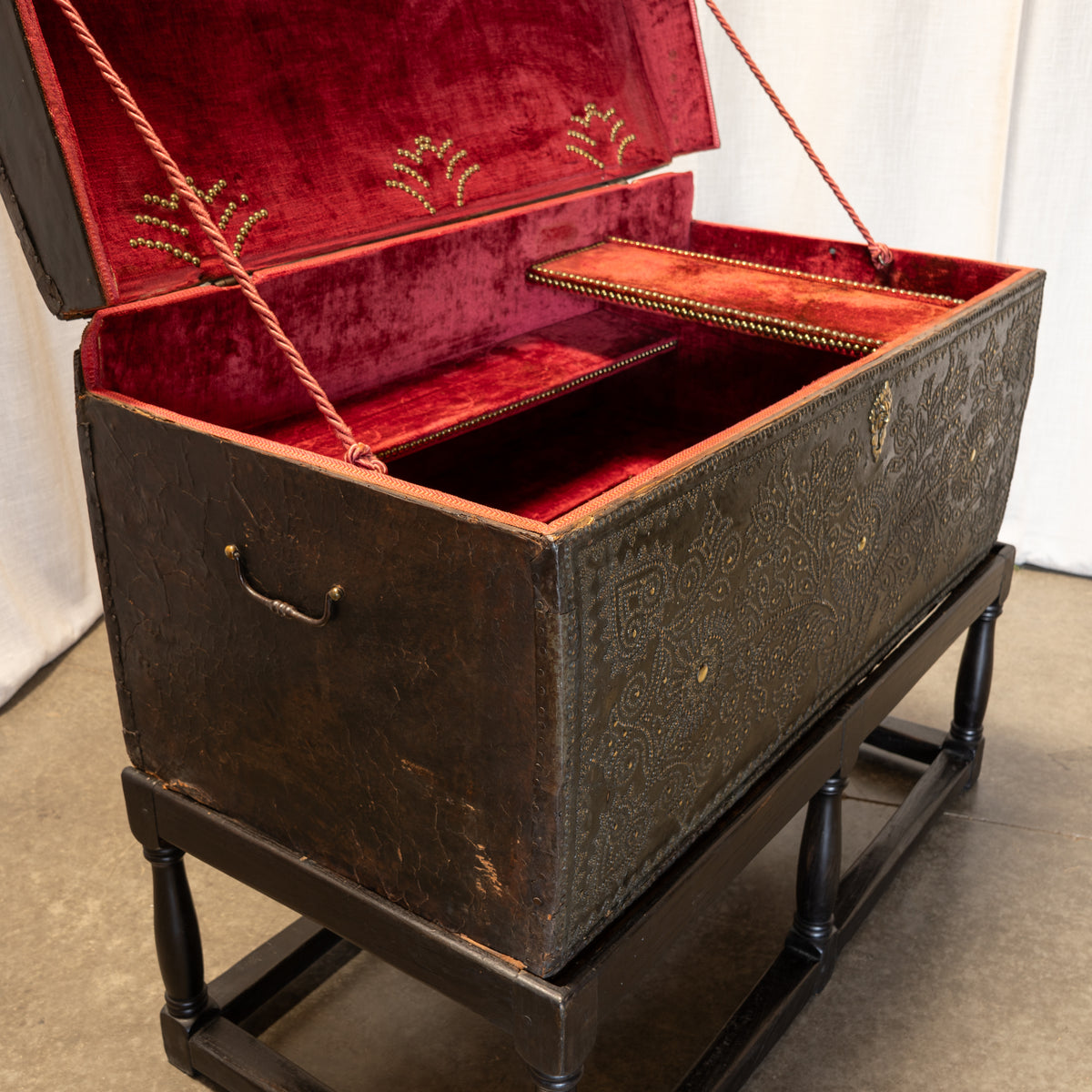 An antique dark brown carved wooden trunk with a deep red velvet-lined interior, displayed open on a black wooden stand.