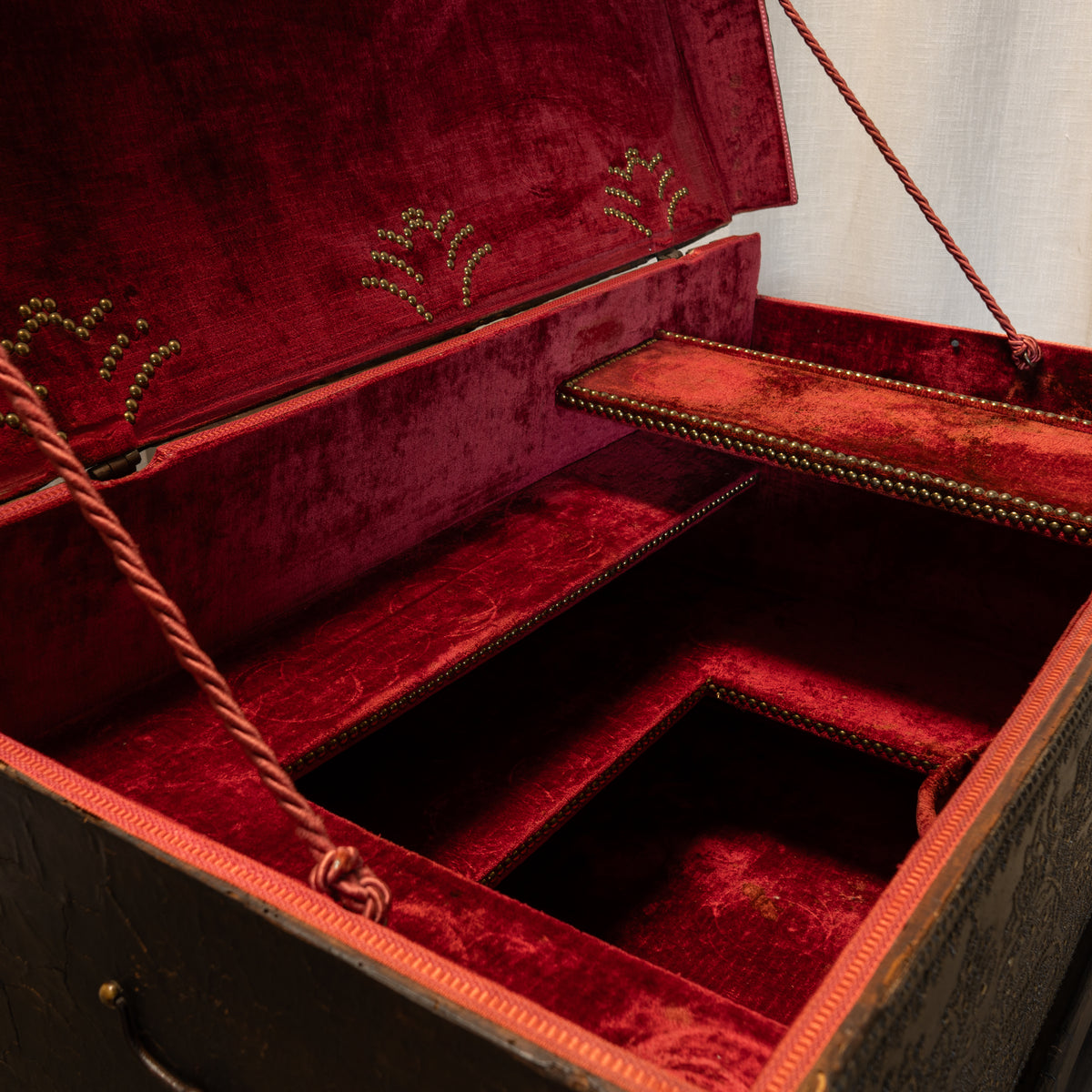 An open vintage trunk or storage box with deep red velvet interior lining, decorative gold beadwork on the lid, and a dark wood exterior with brass hardware.