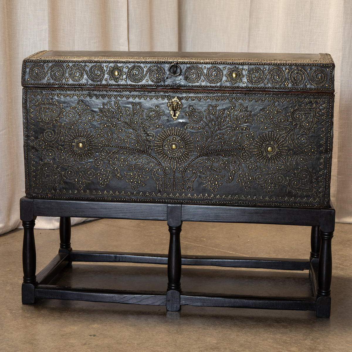 An ornate rectangular metal chest with embossed floral and scrollwork detailing, resting on a dark wooden stand against a beige fabric backdrop.