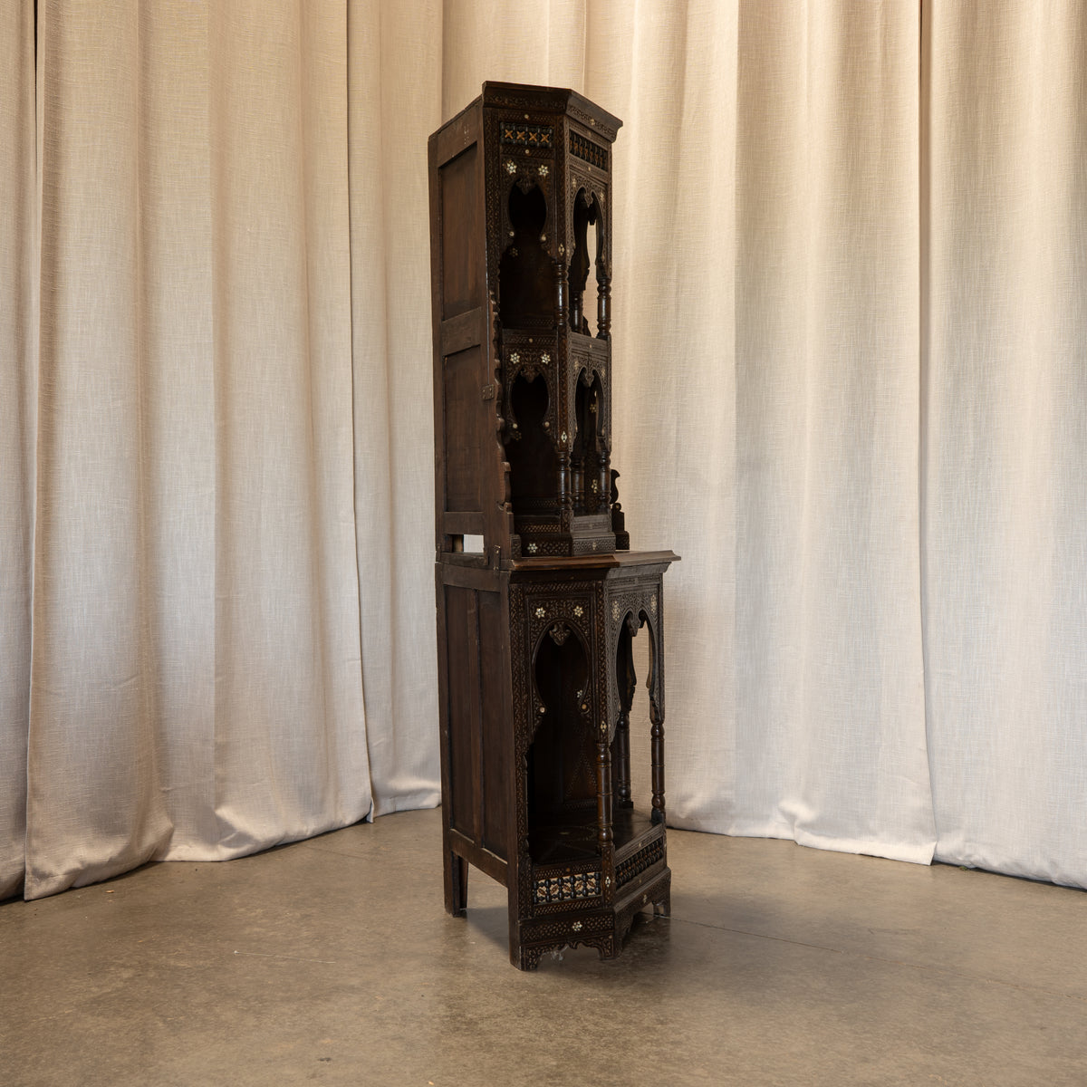 A tall, dark carved wooden display cabinet or hanger stand with ornate open shelving and decorative metal accents, photographed against draped cream fabric curtains and a concrete floor.