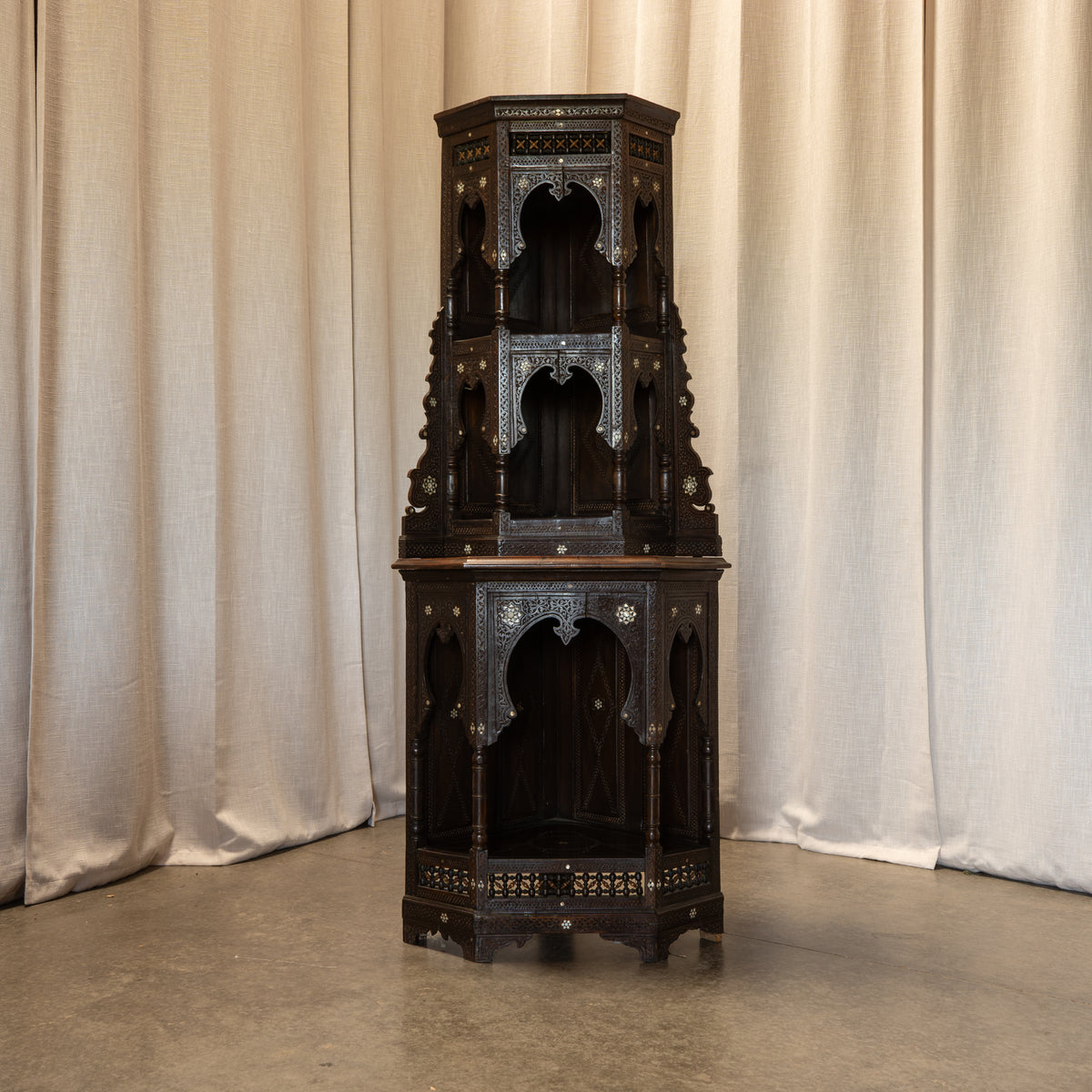 An ornate dark wood Moroccan-style cabinet with intricate brass inlay and arched niche details, set against a backdrop of beige fabric curtains.
