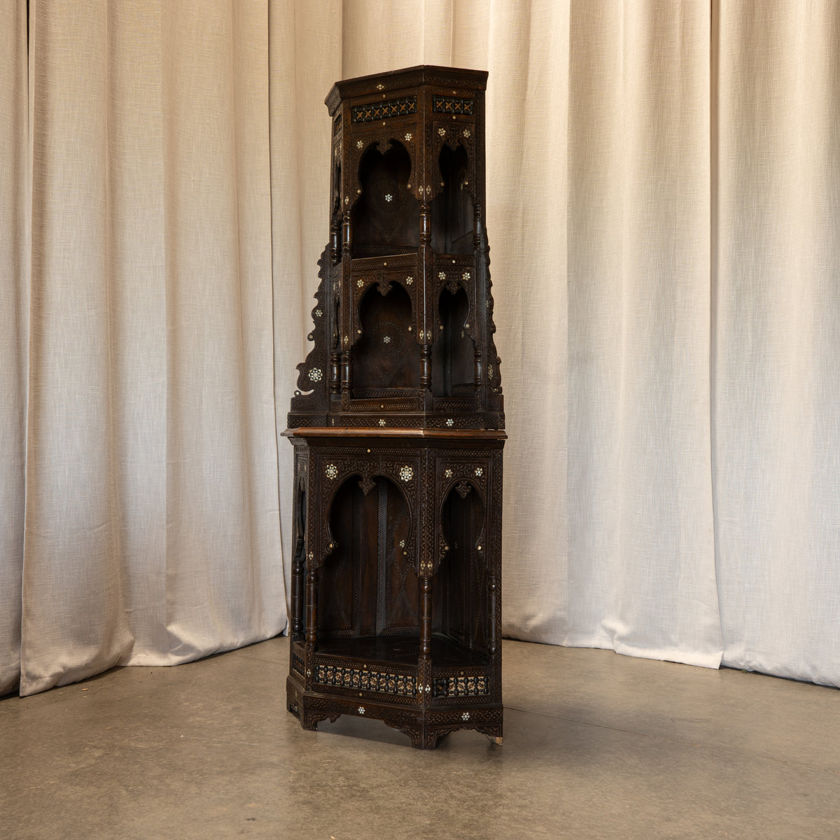 A tall, dark carved wooden corner cabinet with multiple arched niche shelves and brass hardware, photographed against draped cream fabric and a concrete floor.