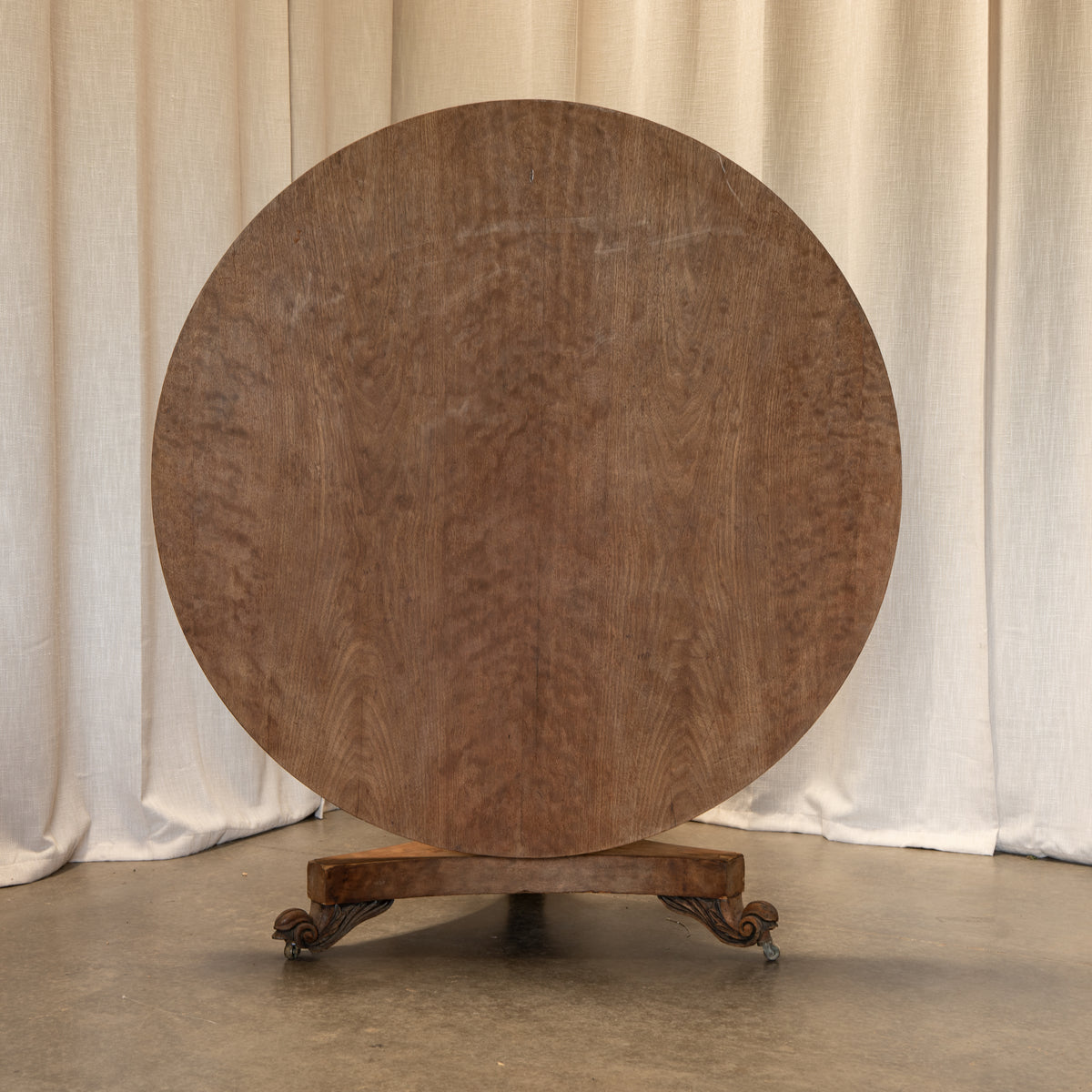 A round wooden table top with a rich brown grain, displayed on a carved wooden stand against a neutral draped fabric backdrop.
