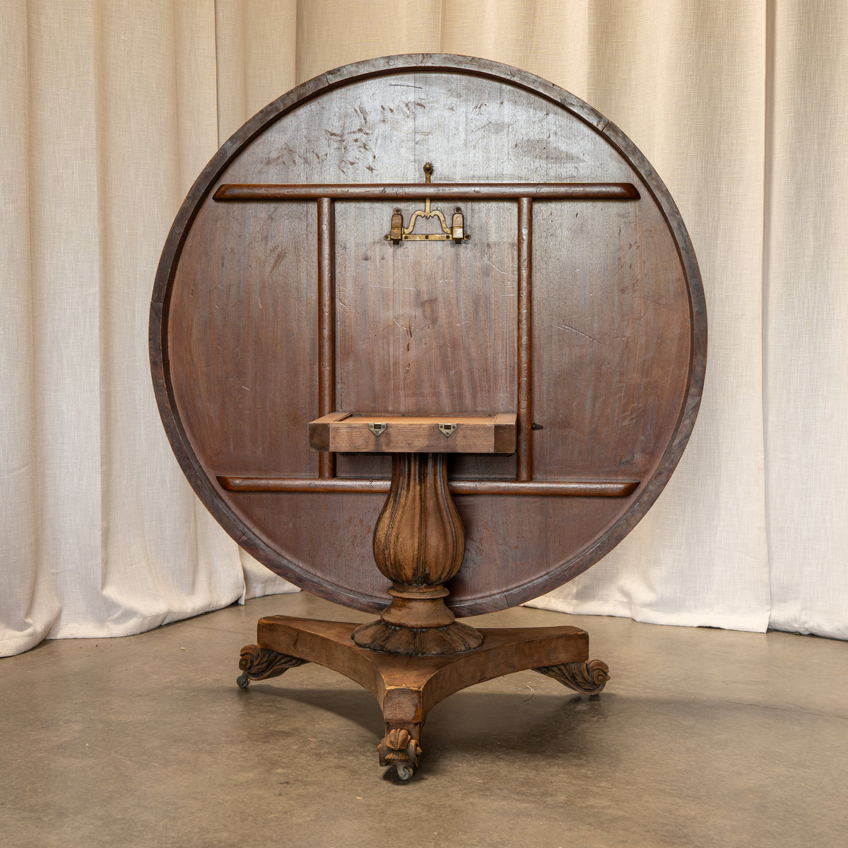 An antique round wooden table with a dark brown finish, featuring a central pedestal base with casters and a small wooden shelf or stand on the underside, photographed against a draped beige fabric backdrop.