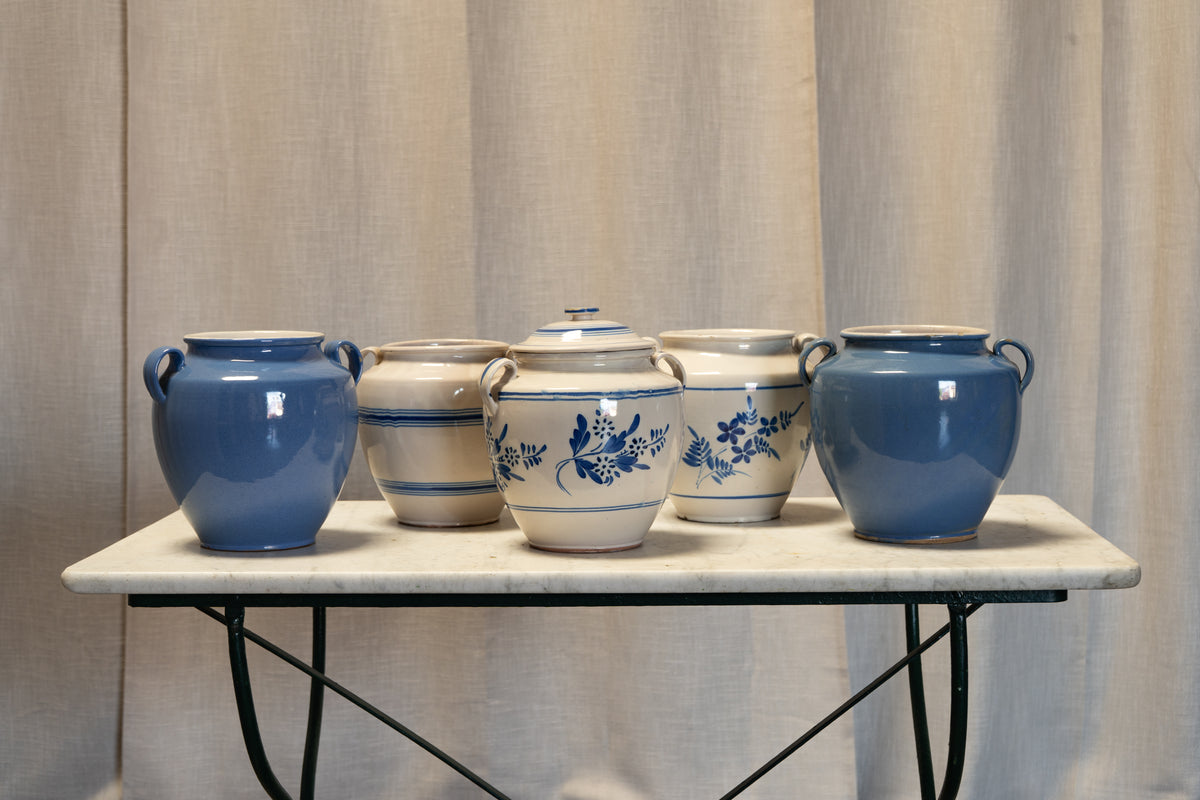 Five ceramic storage jars arranged on a small table against a beige curtain backdrop. Four jars are blue with white handles and floral decoration, and one is cream with blue stripes.