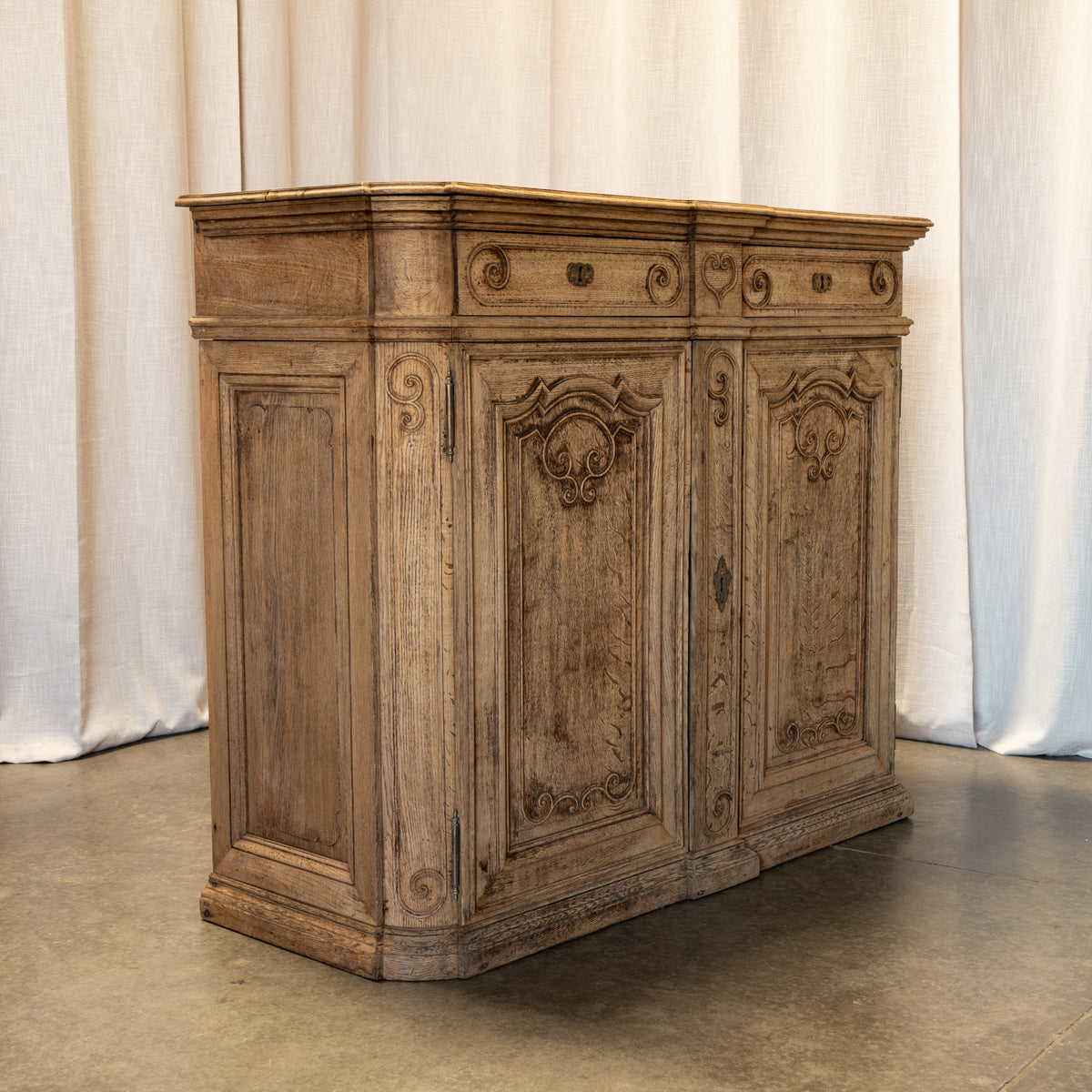 An antique carved wooden cabinet with distressed brown and beige weathered finish, set against a neutral draped fabric backdrop.
