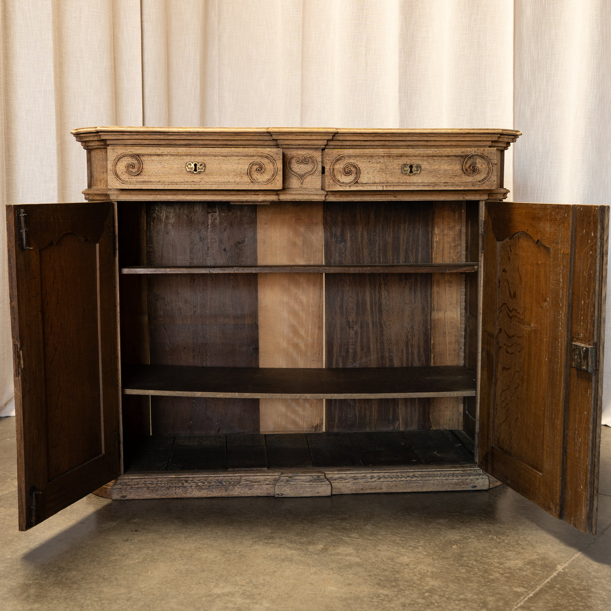 An antique wooden side cabinet with two doors open, showing a rustic dark wood interior and a lighter carved top panel with scroll details, set against a neutral draped fabric backdrop.