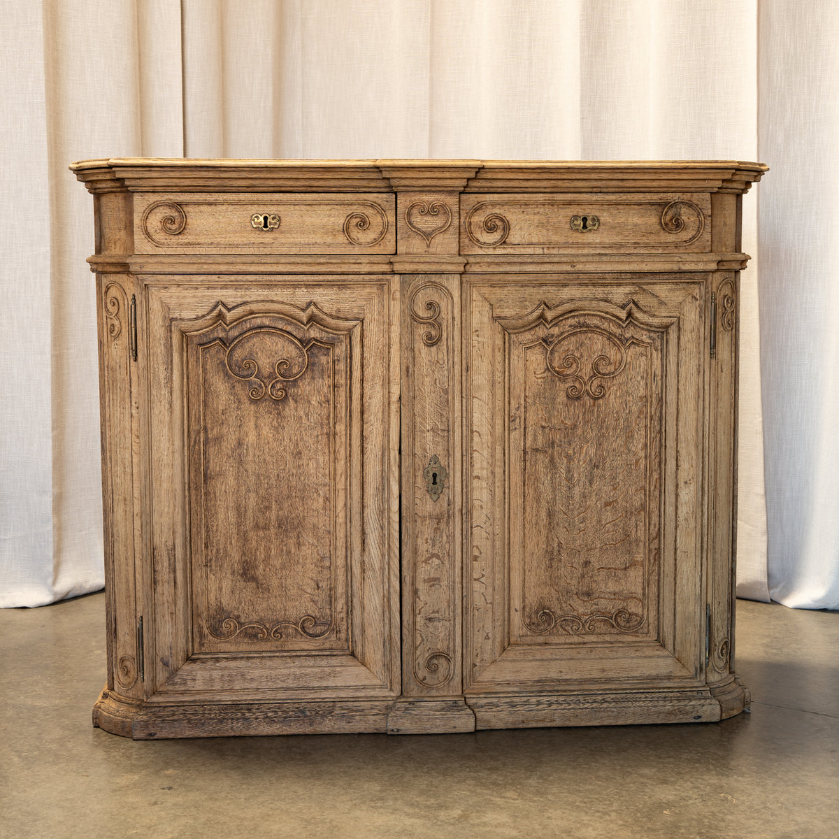 An antique carved wooden cabinet with two doors and a decorative top, shown against a neutral draped fabric backdrop.