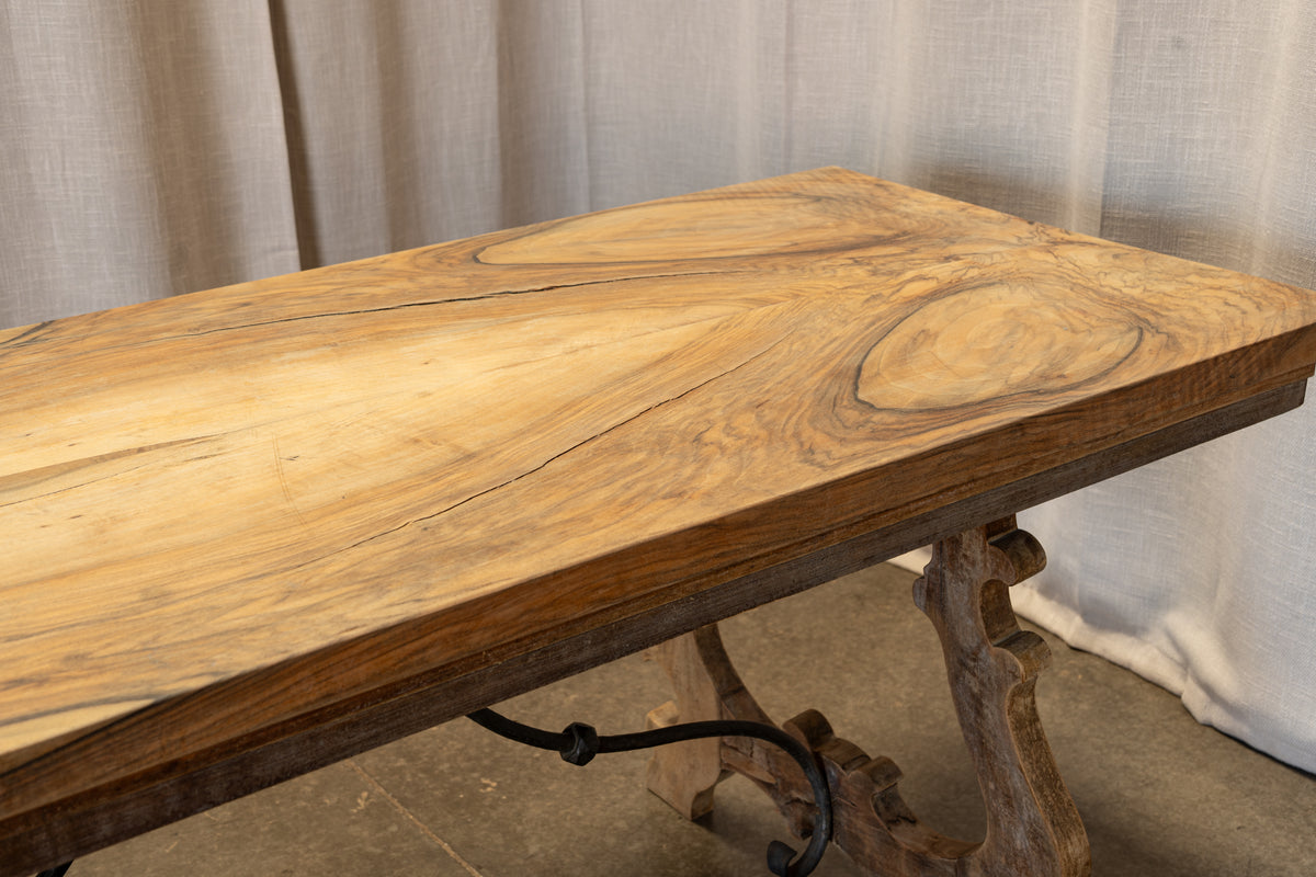 A rustic live-edge wooden table top with a rich grain pattern, mounted on a carved wooden trestle base with black metal scrollwork, set against a neutral draped fabric backdrop.