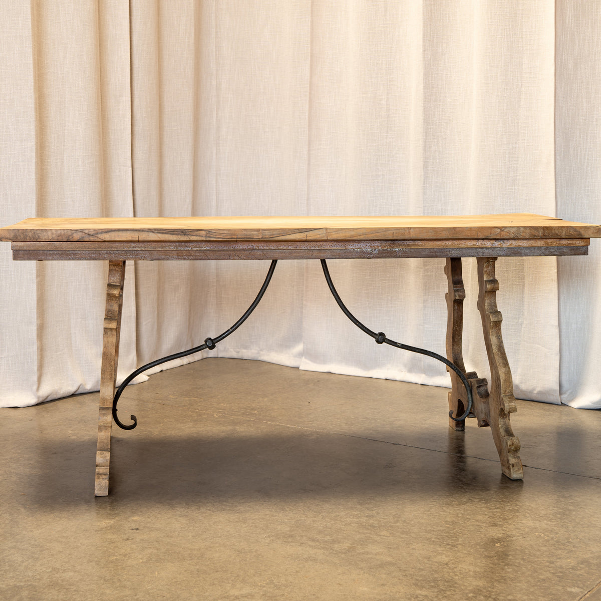 A rustic rectangular wooden table with a weathered light brown top and distressed gray-brown legs, supported by a black metal scroll bracket base, set against a backdrop of draped beige fabric and a concrete floor.