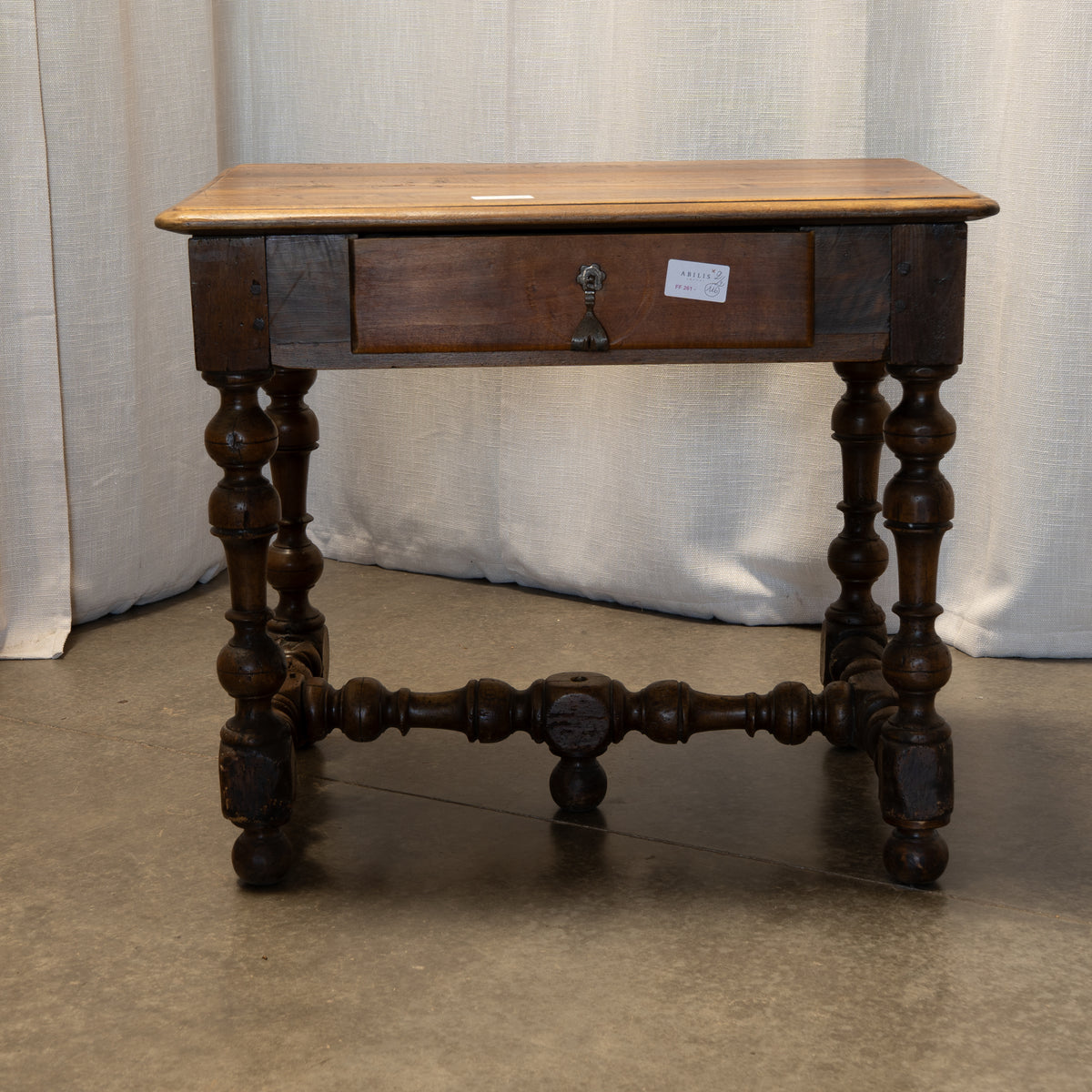 An antique dark wood writing desk with a light wood top, a single drawer with a brass pull, and turned spindle legs, photographed against a white curtain backdrop.