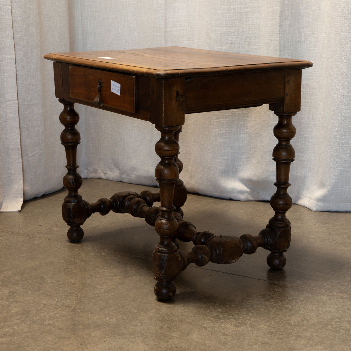A dark brown wooden end table with a single drawer, turned spindle legs, and a decorative stretcher bar, photographed against a draped white fabric backdrop.