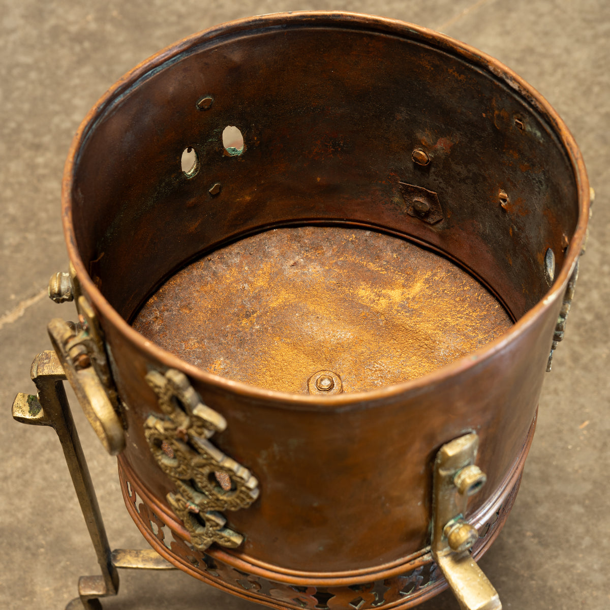 A close-up of a vintage copper radiator or boiler component with brass fittings and a rusted interior, resting on a concrete floor.
