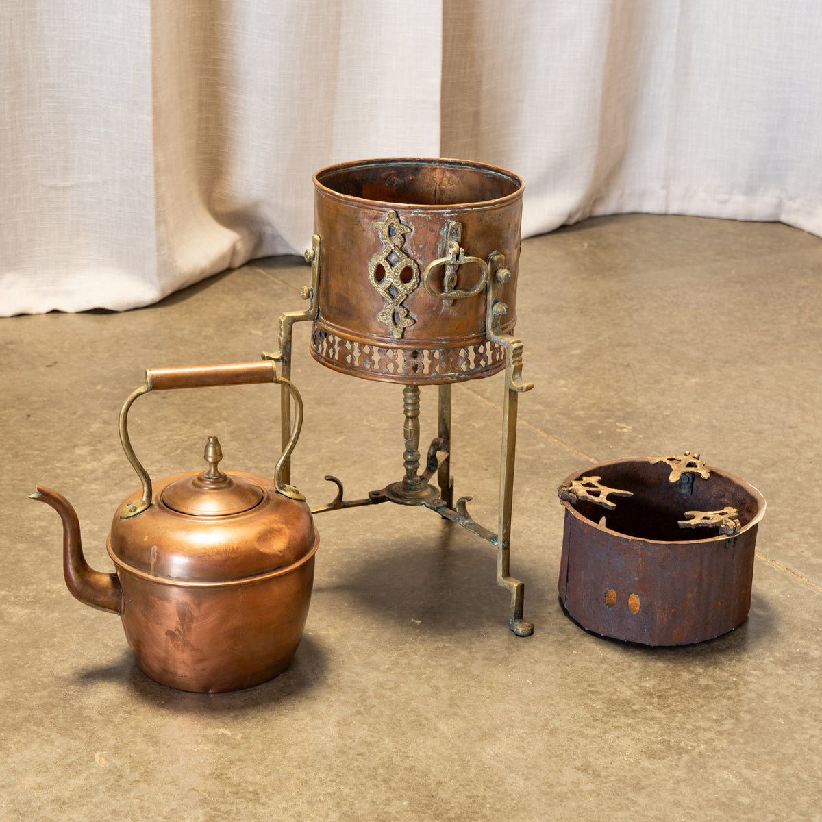 A vintage copper teapot with a wooden handle sits beside a matching copper burner stand and a small copper pot, all displayed on a concrete floor in front of draped fabric.