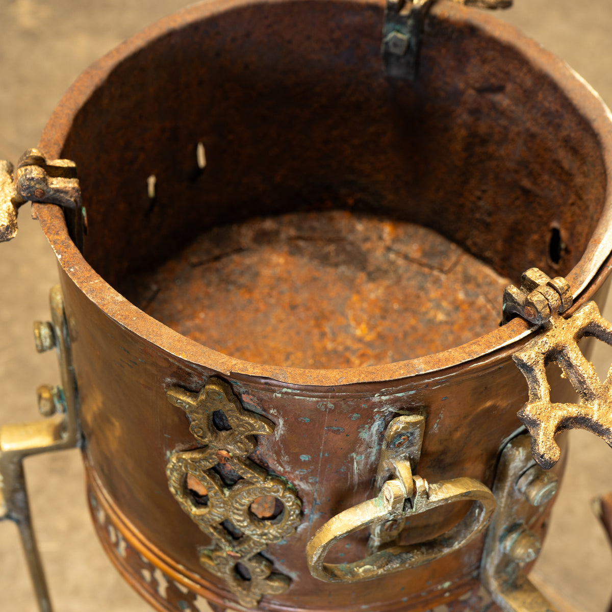 Close-up of a rustic metal container or vessel with a heavily rusted brown exterior, aged bronze-toned hardware, and decorative scrollwork on a neutral concrete floor.