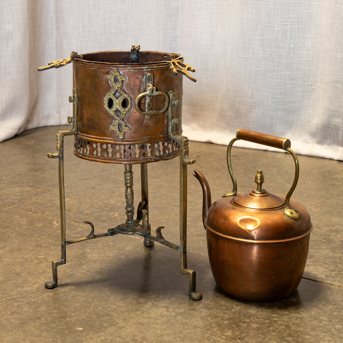 A vintage copper tea or coffee pot on a decorative metal stand, shown next to a matching copper kettle against a draped fabric backdrop.