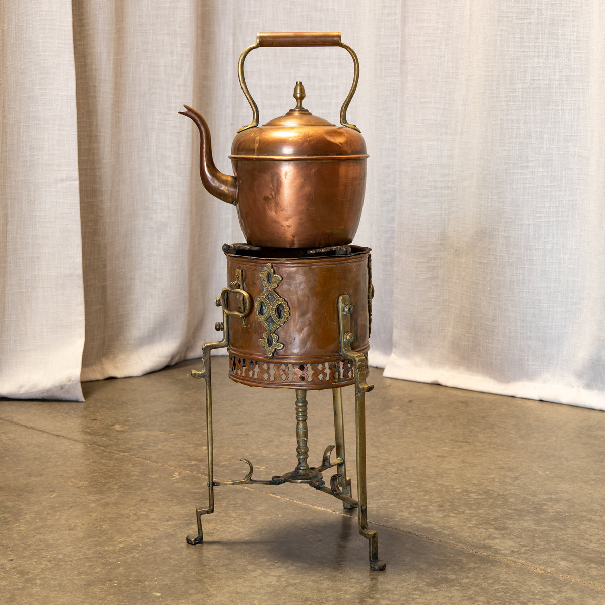 A vintage copper teapot with a wooden handle sits on top of an ornate matching copper and brass stove, set against a neutral draped fabric backdrop.