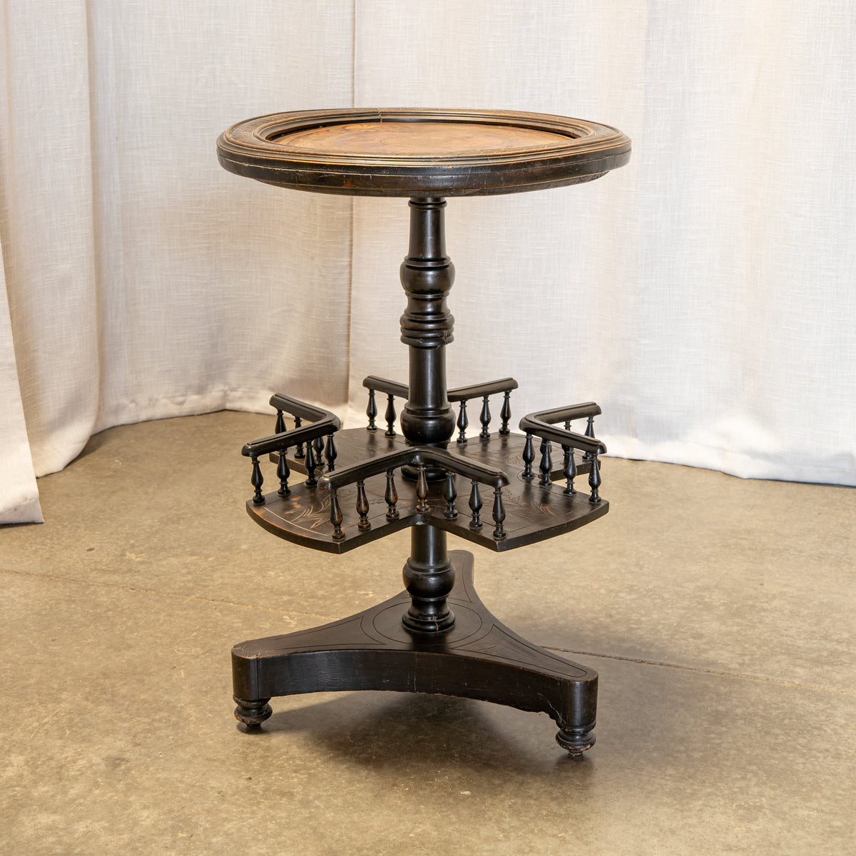 A vintage dark wood pedestal table with a round top, a central turned post, and a three-tiered side-arm shelf section, photographed against a draped fabric backdrop.